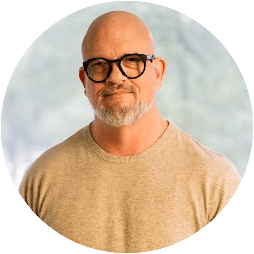 Smiling bald man with black glasses and gray beard wearing a beige sweater, neutral background.