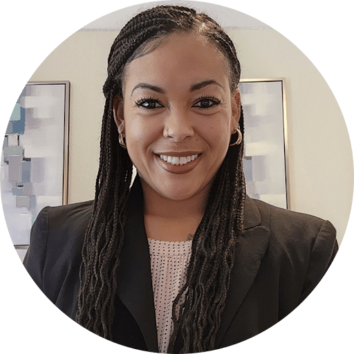 Smiling woman with braided hair wearing a black blazer in an office setting.