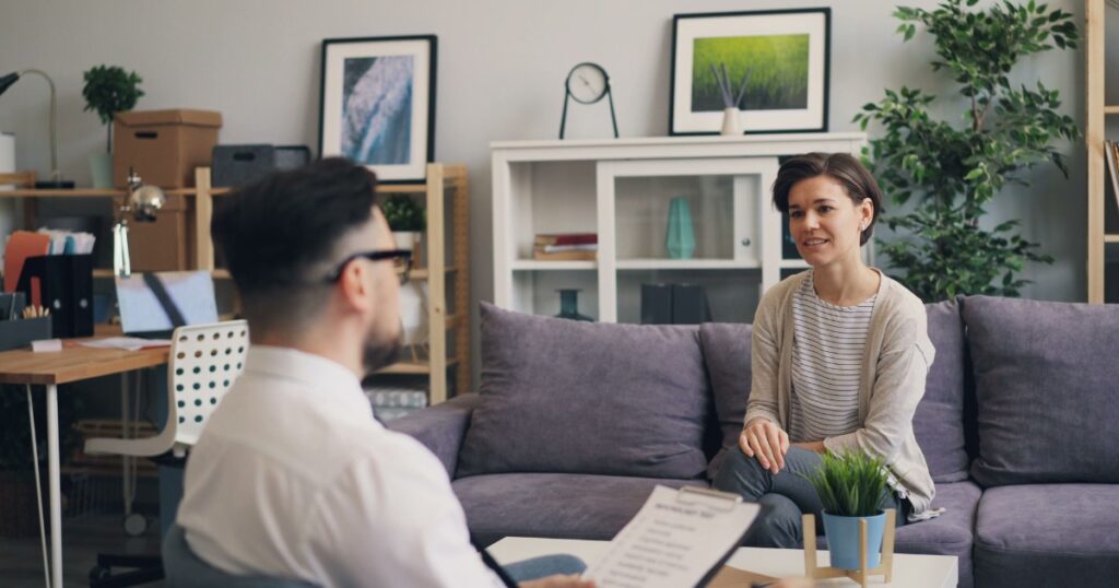 developmental psychology - Treat Mental Health Man with a clipboard and glasses sits facing a woman on a gray sofa in a bright, modern office with plants and shelves in the background.
