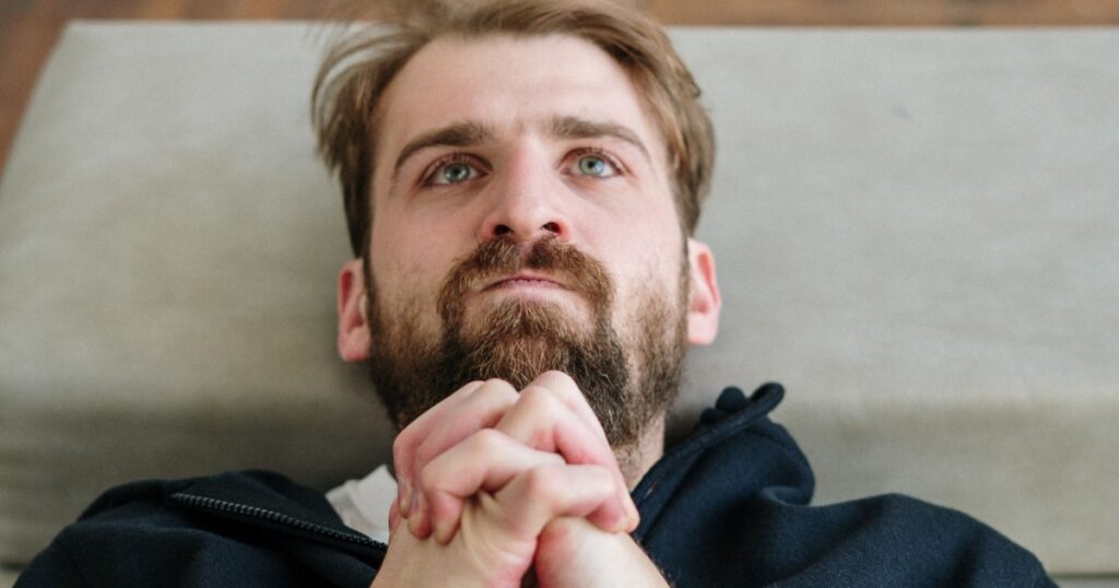 developmental psychology - Treat MH - Treat Mental Health Bearded man lying down with hands clasped beneath his chin, gazing upward as if praying or pondering.