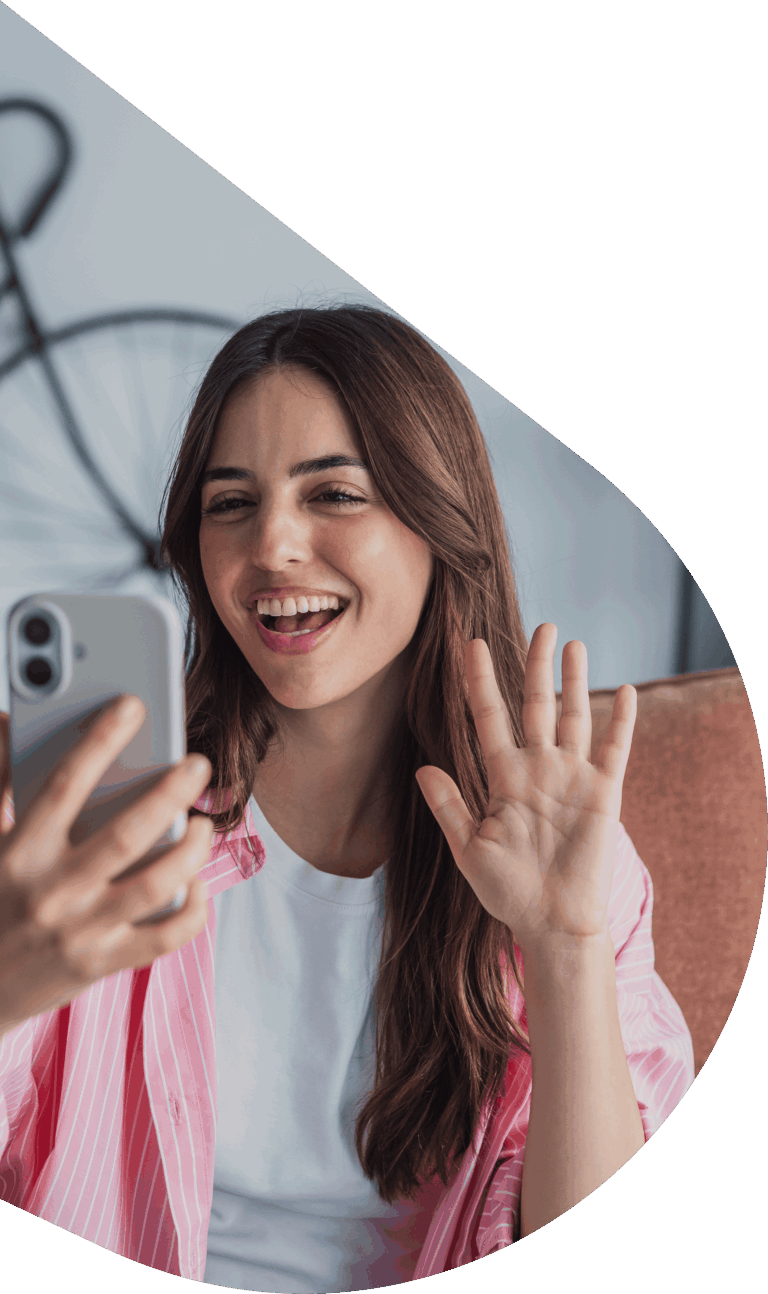 Smiling young woman takes a selfie with her smartphone and waves at the camera, pink striped shirt visible.